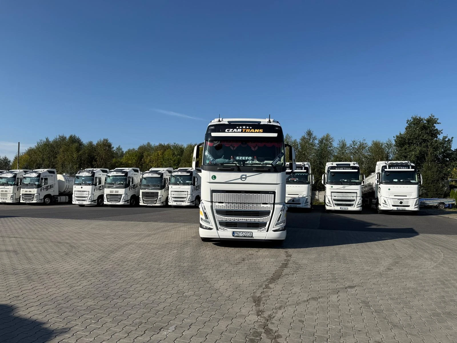 Biały ciągnik Volvo CZAR-TRANS z rzędem firmowych pojazdów w tle, na placu firmy